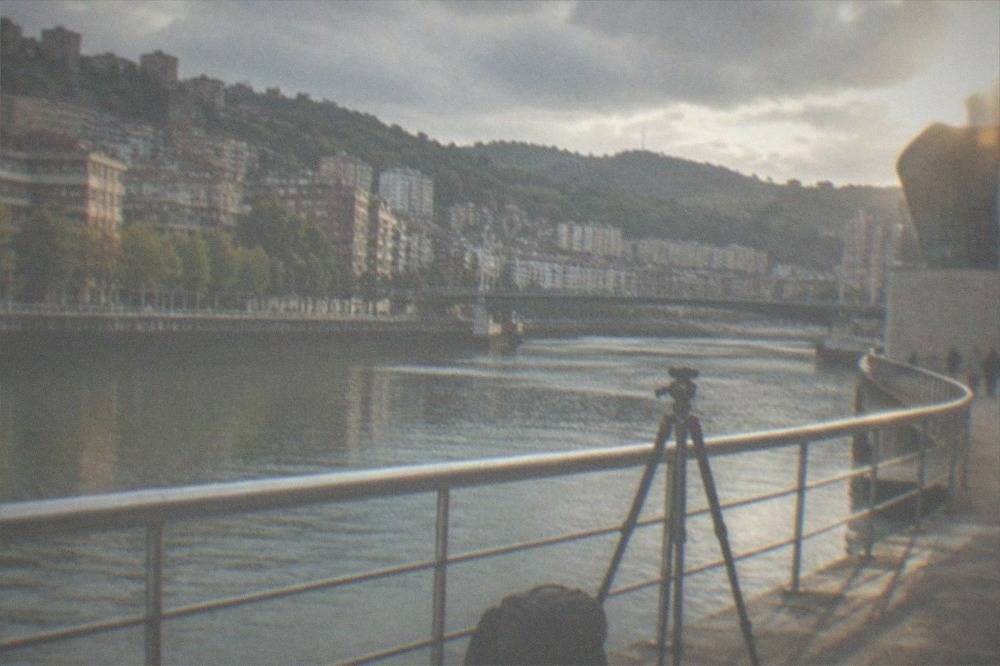 A photographer's camera bag and tripod on a stone riverside promenade beside a modern museum building