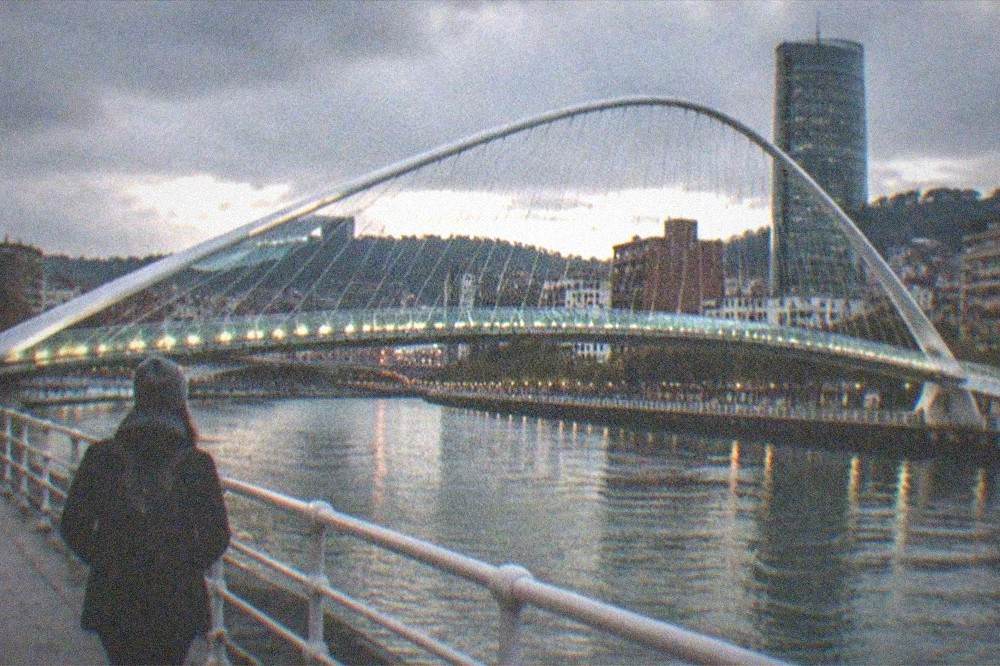 A solo traveler at dusk on the riverside promenade with the illuminated Zubizuri bridge and Bilbao skyline behind
