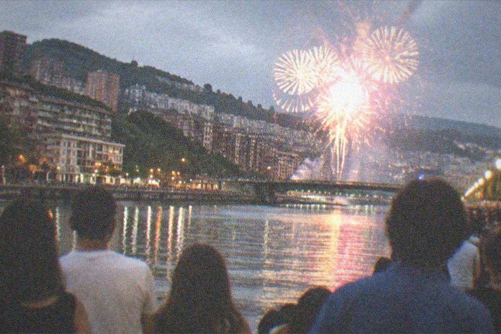 A crowd watching fireworks over a river in a city at night