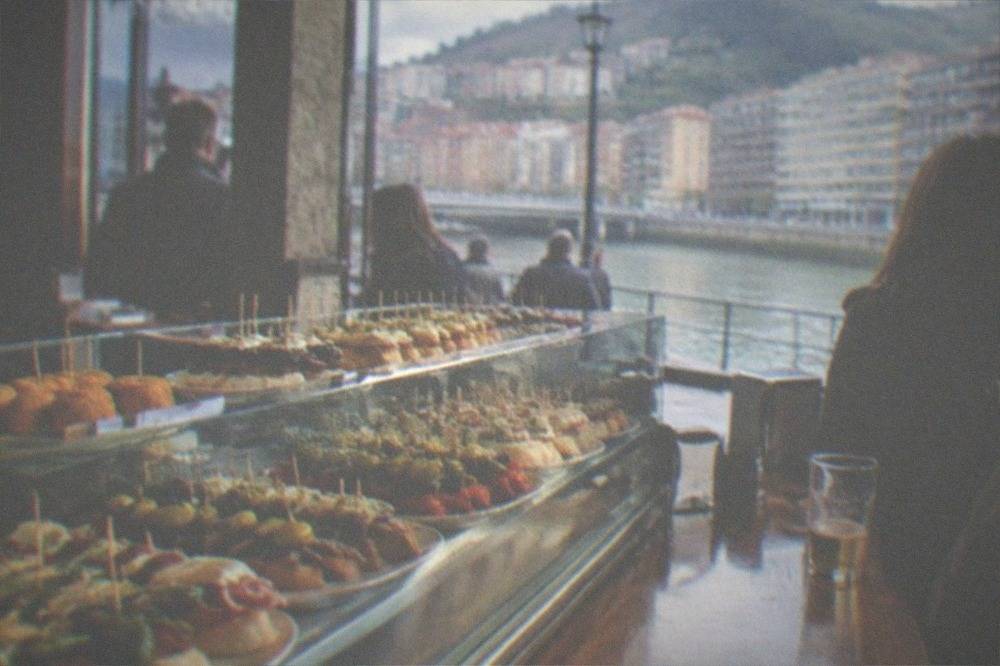 A crowded pintxos bar counter in Bilbao with small skewered snacks displayed under glass