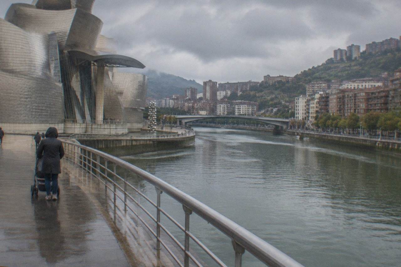 A parent pushing a stroller along a riverside walkway beside a curved titanium museum building in Bilbao