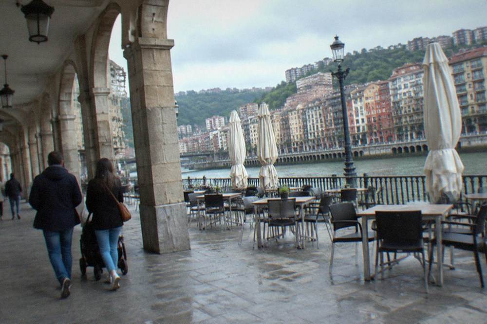A family with a stroller walking through an arcaded square with cafe tables in Bilbao