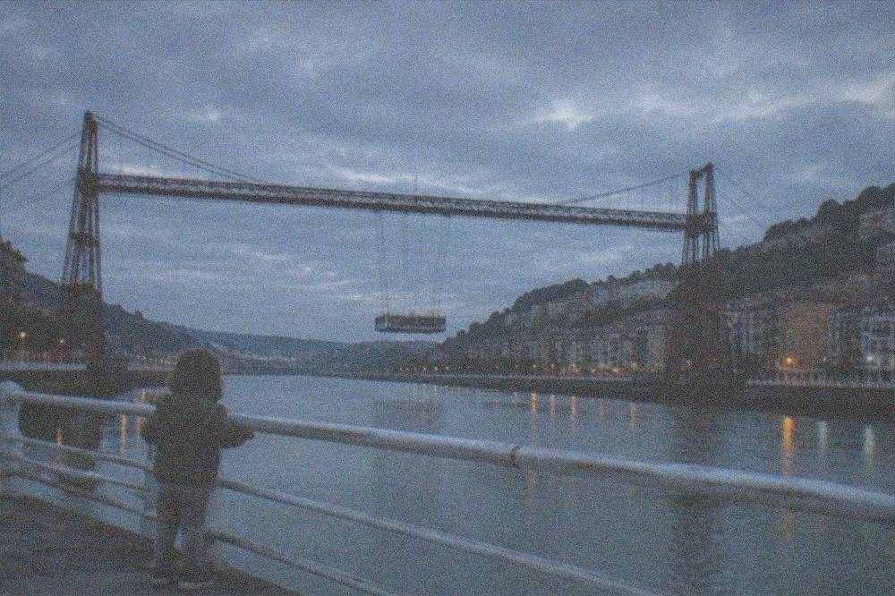 A toddler watching a suspended gondola bridge crossing a river between two towns