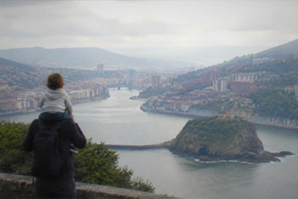 A parent carrying a toddler on their shoulders looking out over a rocky coastline and a small island hermitage