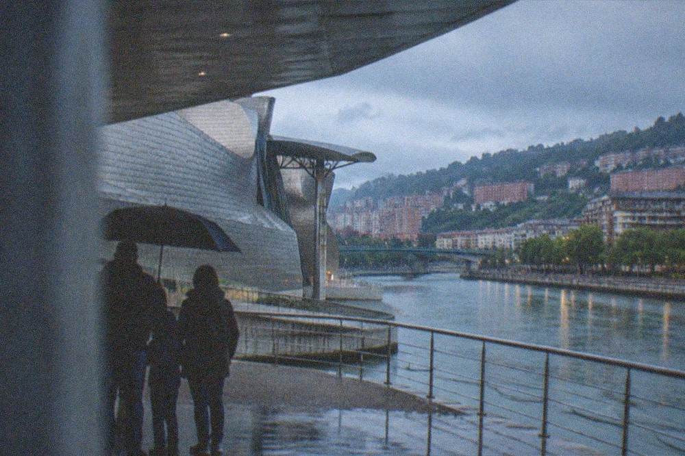 A family sheltering under a wide museum canopy while rain falls on a titanium-clad building