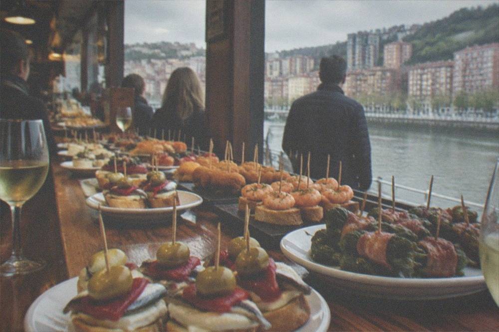 A crowded pintxos bar counter in Bilbao loaded with small skewered bites and glasses of txakoli wine