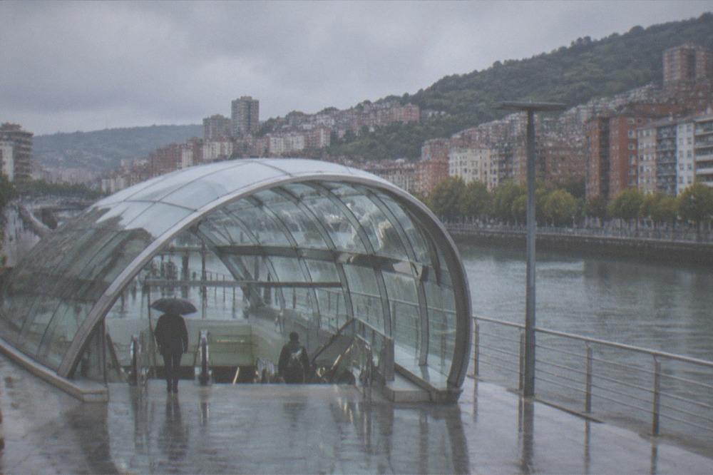 A sleek Bilbao metro station interior with a Foster-designed glass canopy entrance above ground in the rain