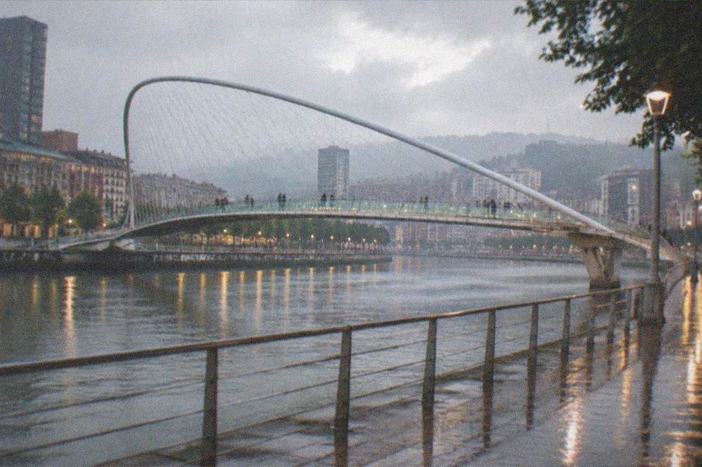 An evening view of Bilbao's illuminated Zubizuri bridge and riverside walkways reflected in wet pavement