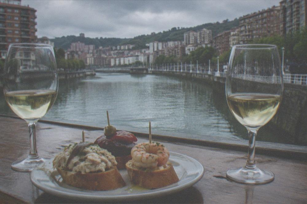 A plate of pintxos and glasses of txakoli wine on a wooden bar top